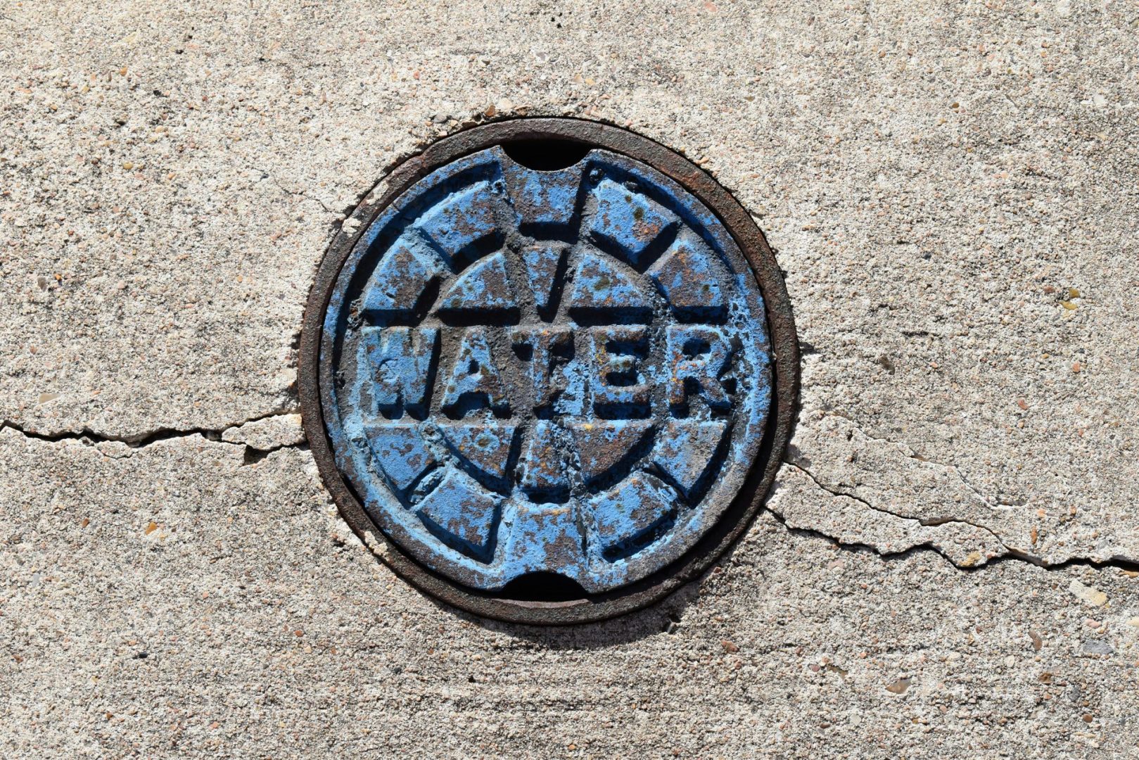 Blue round manhole cover embedded in rough concrete, with rusted rim and peeling paint.